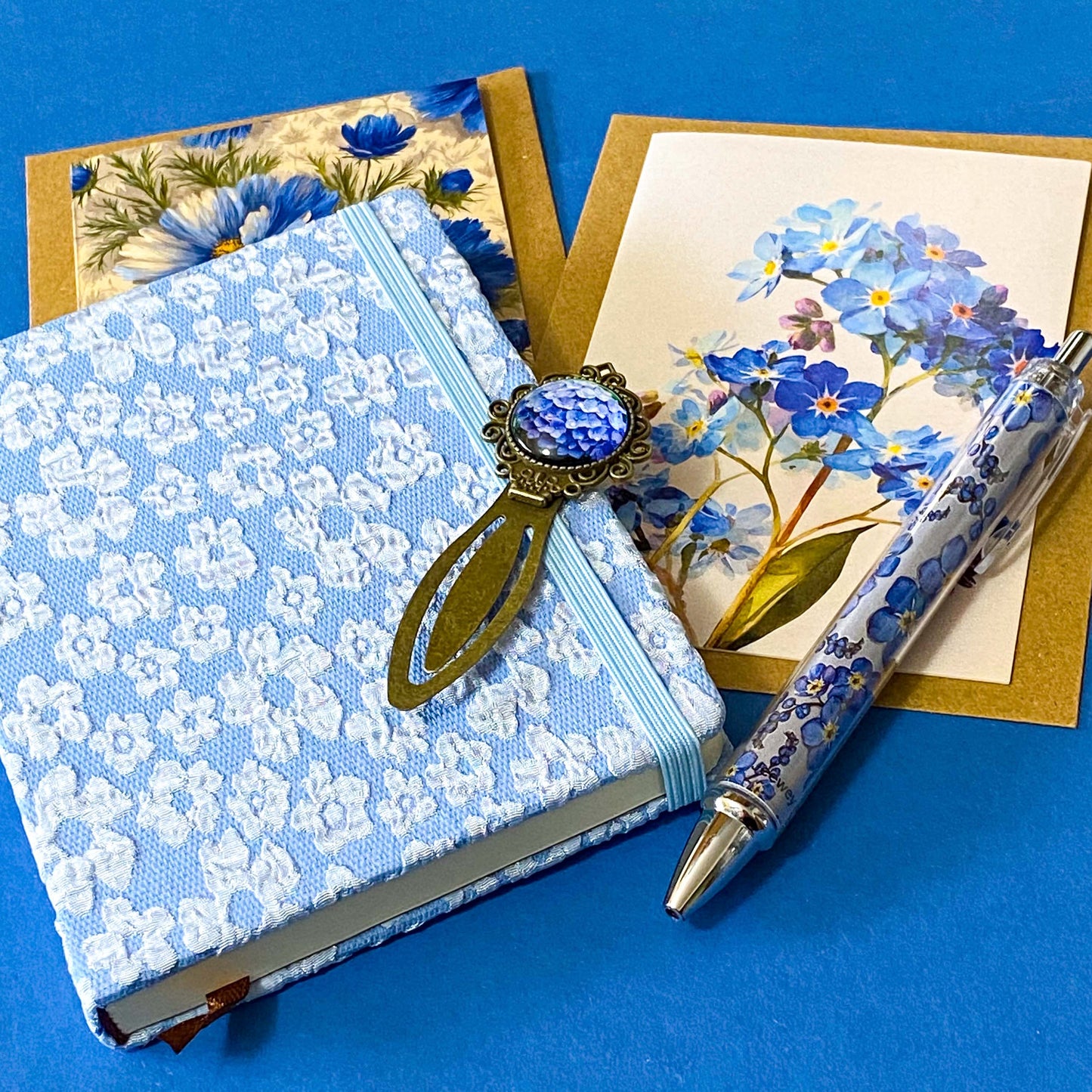 soft blue floral letterbox writing set featuring a textured hardback blue and white floral notebook, matching pen, forget me not cabochon bookmark and blank greetings cards and kraft envelope. uk gift shop.
