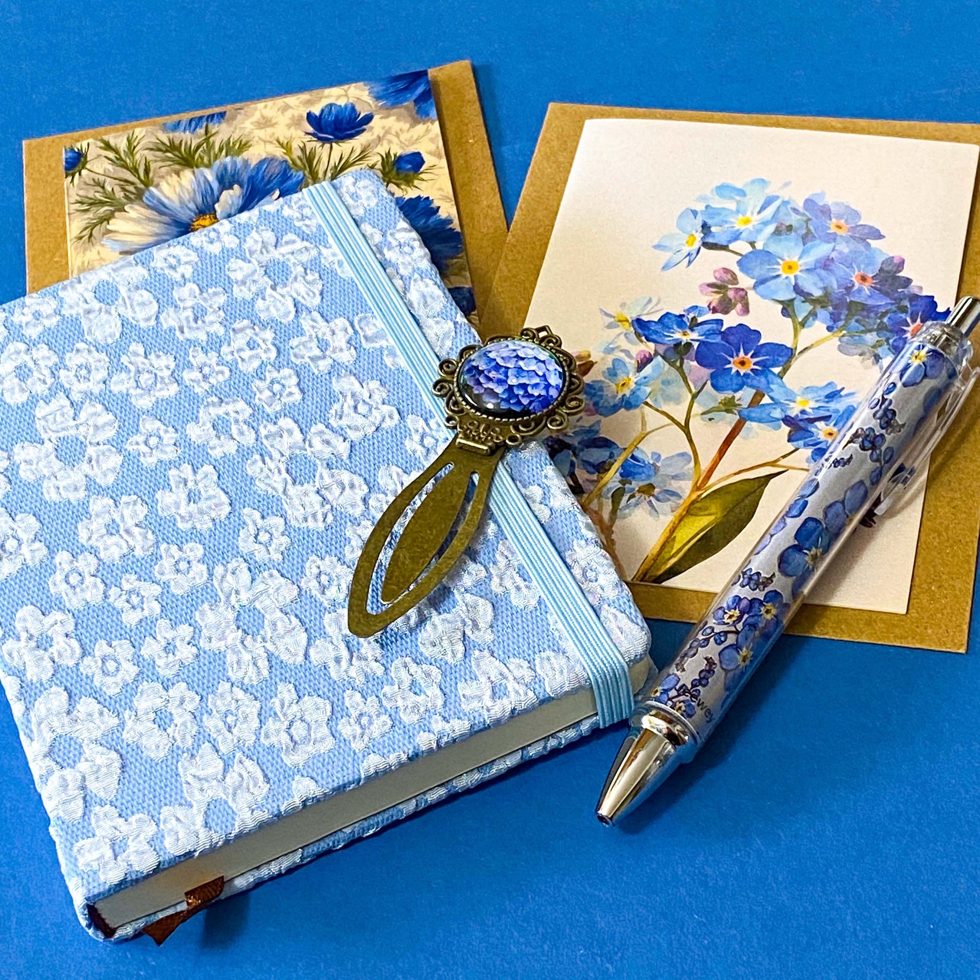 soft blue floral letterbox writing set featuring a textured hardback blue and white floral notebook, matching pen, forget me not cabochon bookmark and blank greetings cards and kraft envelope. uk gift shop.