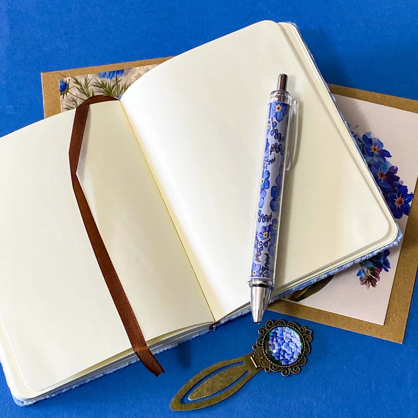 soft blue floral letterbox writing set featuring a textured hardback blue and white floral notebook, matching pen, forget me not cabochon bookmark and blank greetings card and kraft envelope. uk gift shop.