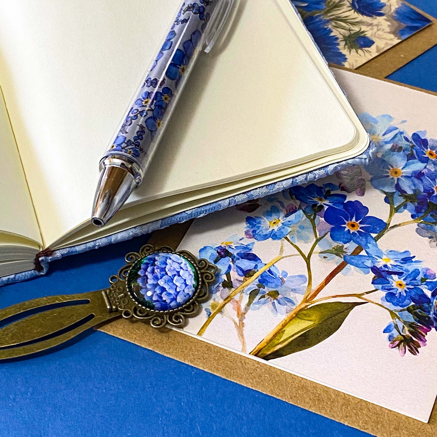soft blue floral letterbox writing set featuring a textured hardback blue and white floral notebook, matching pen, forget me not cabochon bookmark and blank greetings card and kraft envelope. uk gift shop.