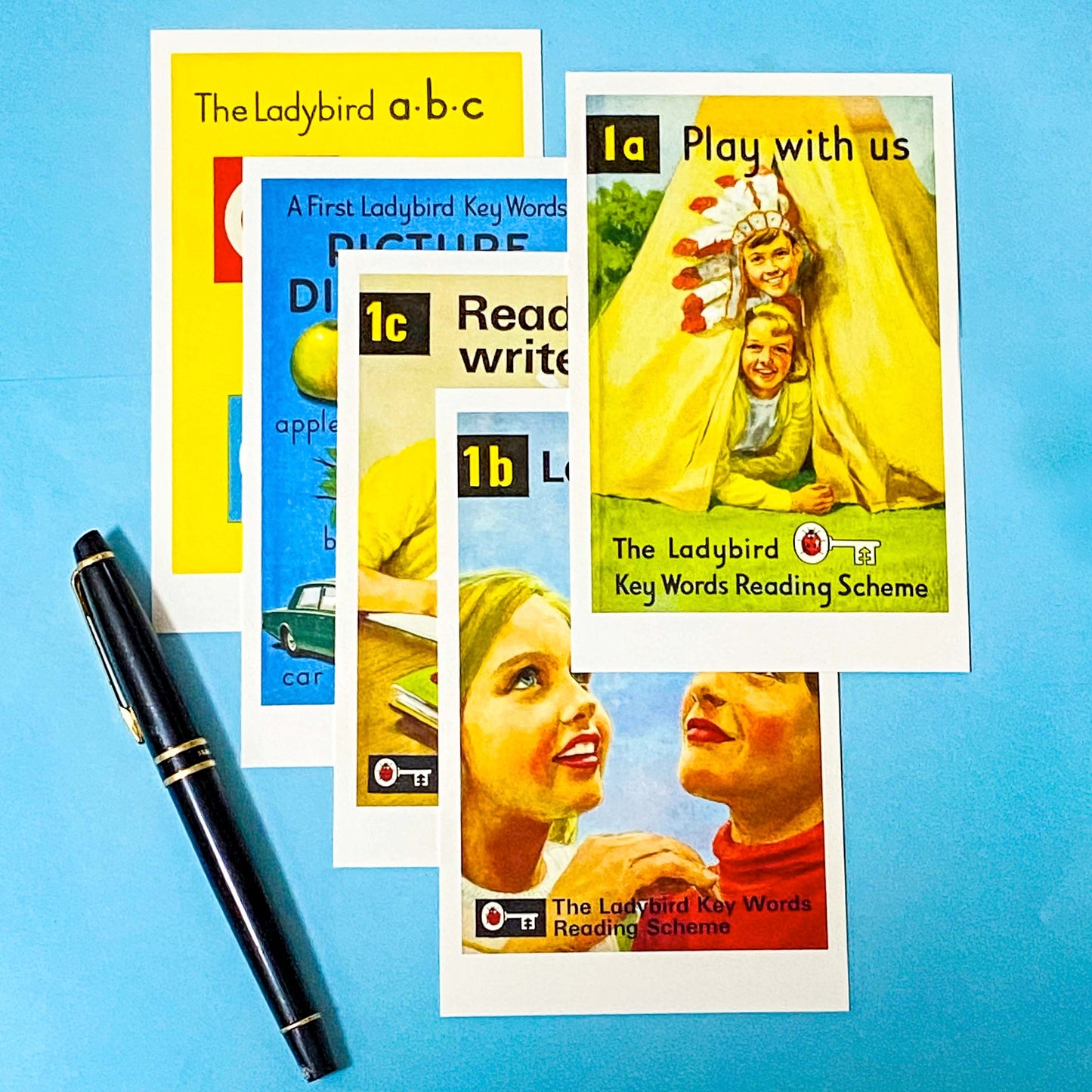 Collection of Ladybird books postcards from the Key Words Reading Scheme on a blue background with a pen.