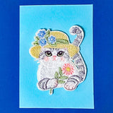 Embroidered patch of a cat applique wearing a flower hat on a blue card
