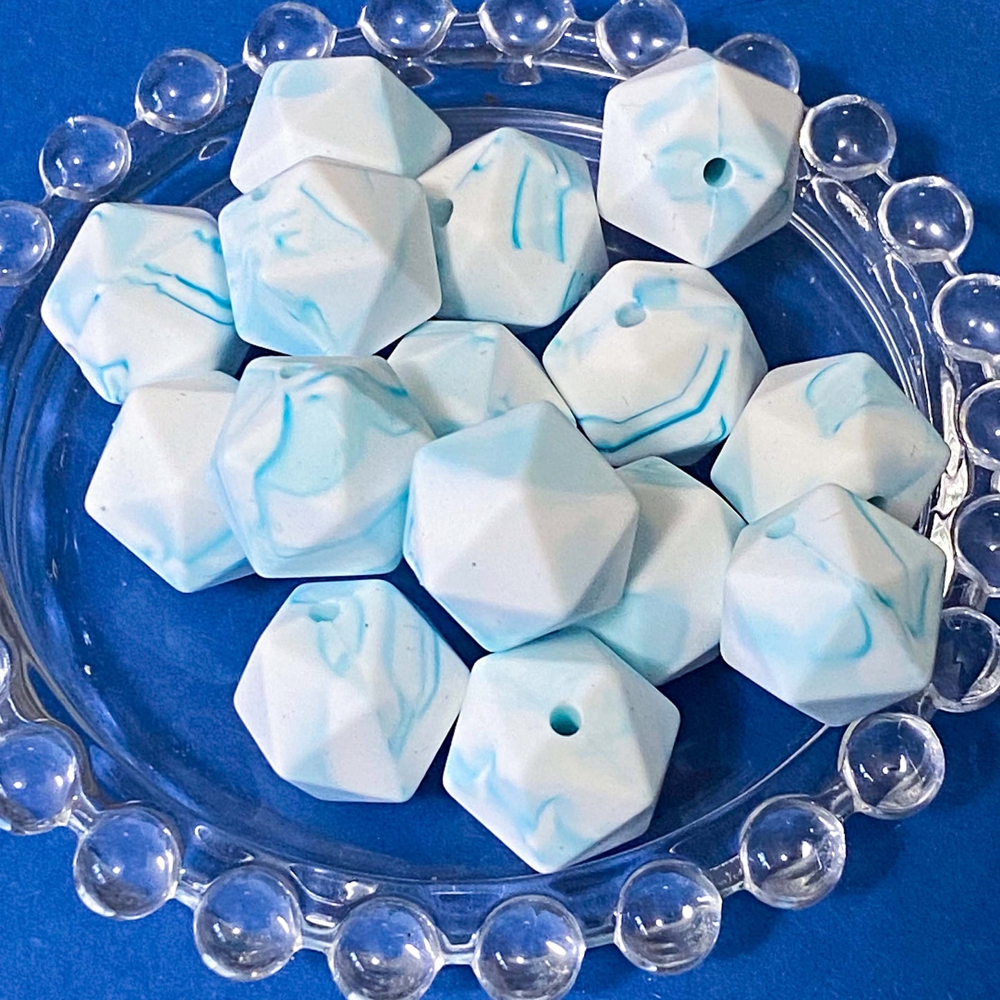 A pile of faceted, dodecahedron-shaped beads with a white and light blue marble swirl pattern, displayed on a clear glass plate against a solid blue background.
