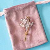 pale pink enamel iris brooch with gold tone metal on a pink velvet drawstring bag on a blue background