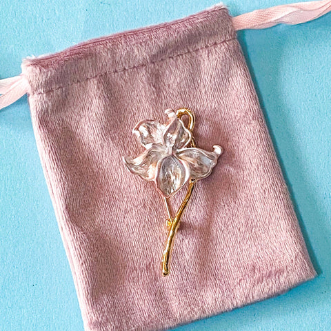 pale pink enamel iris brooch with gold tone metal on a pink velvet drawstring bag on a blue background
