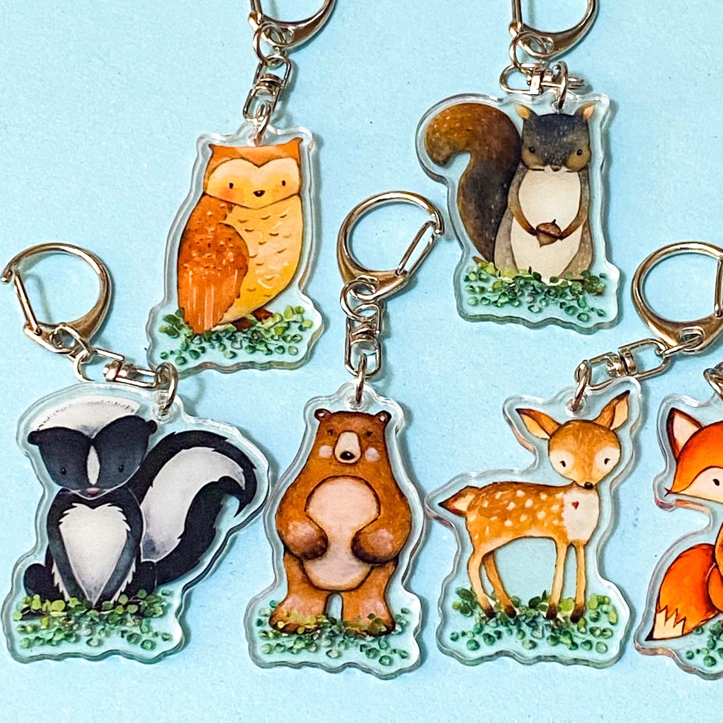 Set of animal keychains including an owl, squirrel, skunk, bear, deer, and fox on a light blue background.