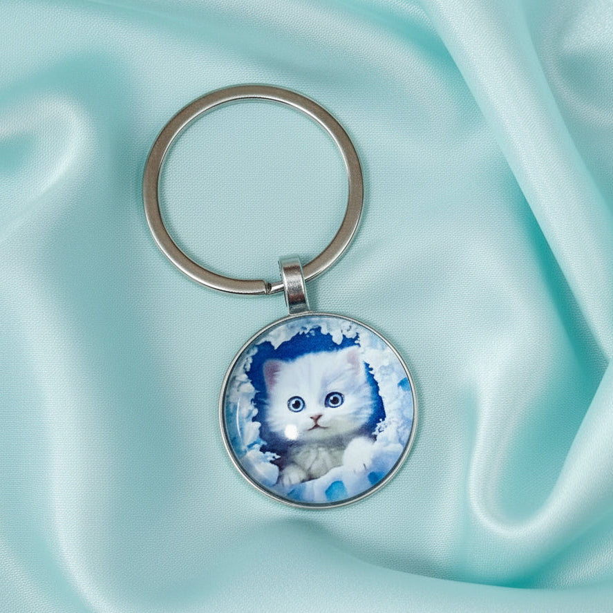 A round metal keychain featuring a printed image of a white kitten in a snowy scene, attached to a silver split ring and photographed on a bright blue background.